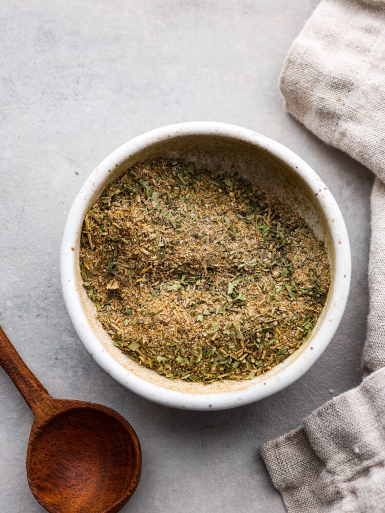 Top view of turkey seasoning in a small white bowl with a wooden spoon and tan kitchen towel next to the bowl.