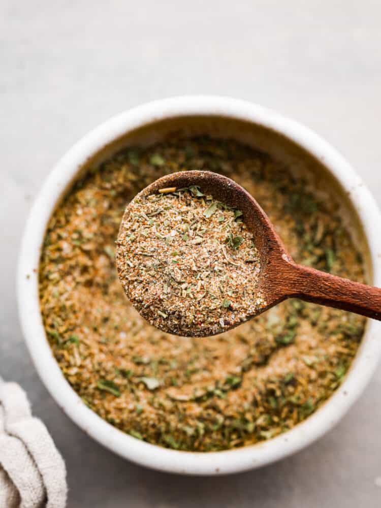 Close up view of a wooden spoon lifting out a spoonful of seasoning. A tan kitchen towel is next to the bowl.