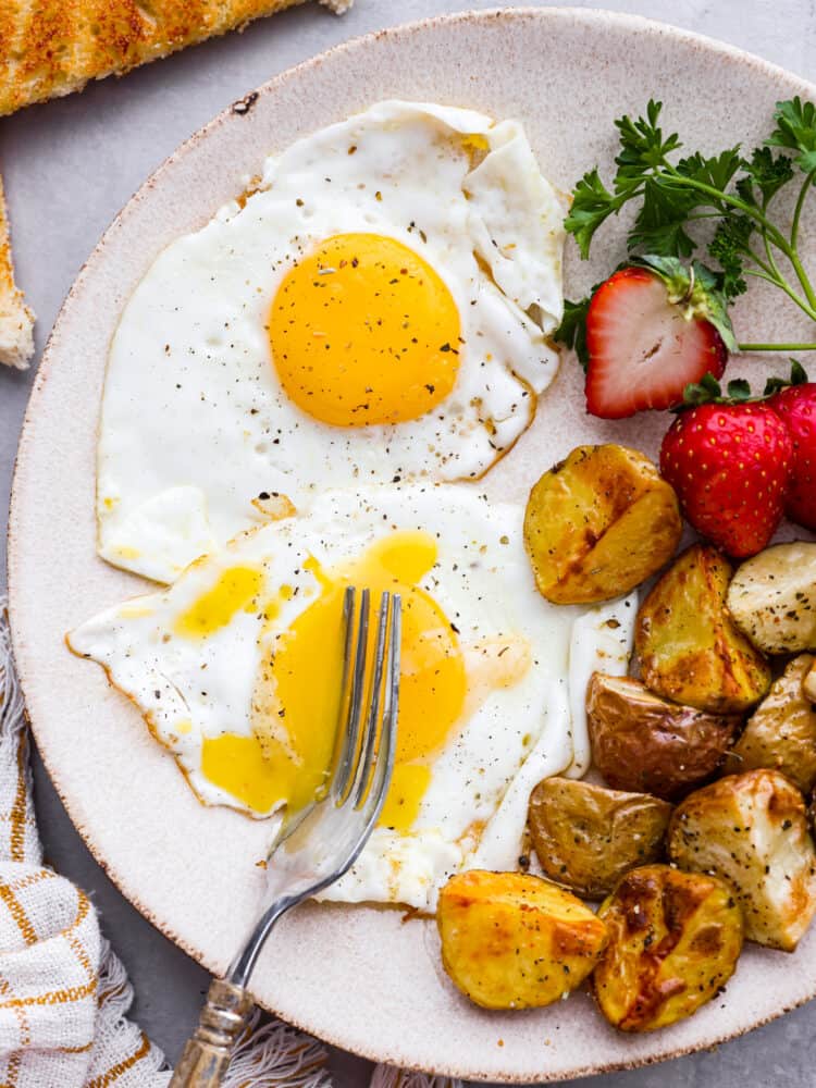 A plate of sunny side up eggs with a fork cutting through it with strawberries and potatoes next to it.