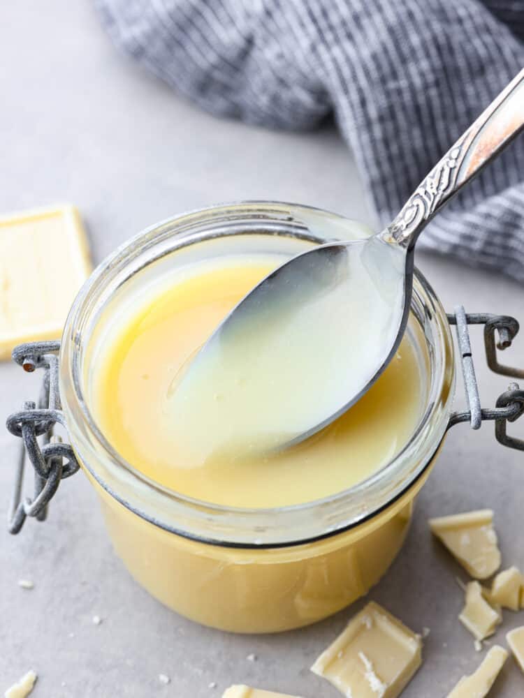 White chocolate sauce in a jar with a silver spoon taking a scoop out with white chocolate pieces around it.