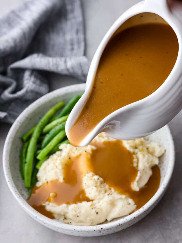 Brown gravy in a sauce pot being poured over some mashed potatoes in a bowl.