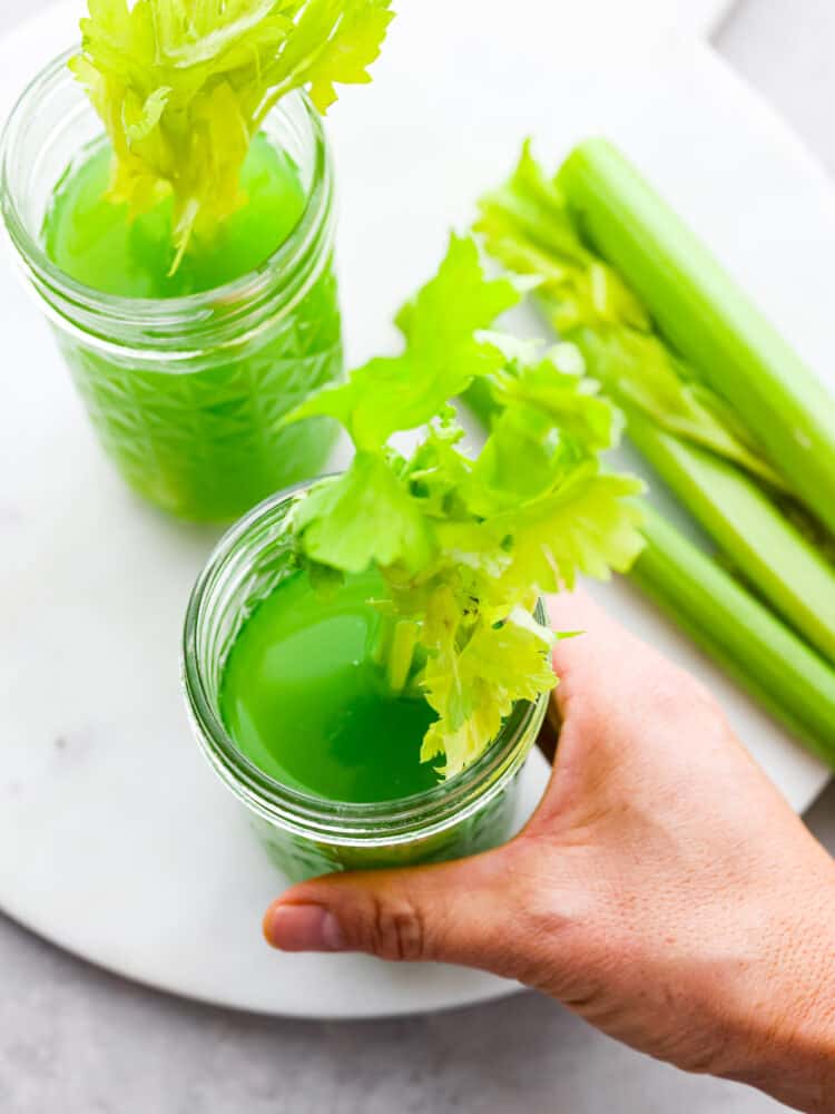 Top view of two glasses of celery juice in mason jars. A leafy celery stalk is styled inside each glass of juice. Celery stalks are styled next to the glasses of juice. A hand is holding one of the glasses.