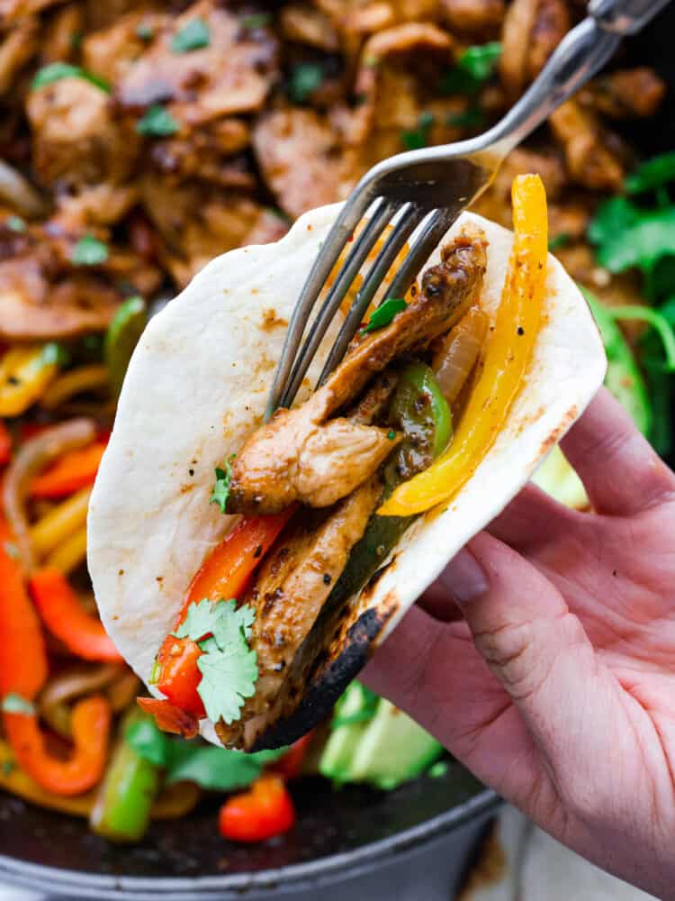 A hand holding a tortilla filling it with chicken and vegetables with a fork.