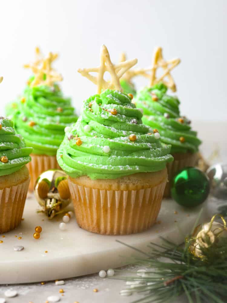 Hero image of Christmas tree cupcakes on a white marble serving board. There are ornaments and a garland surrounding the cupcakes.
