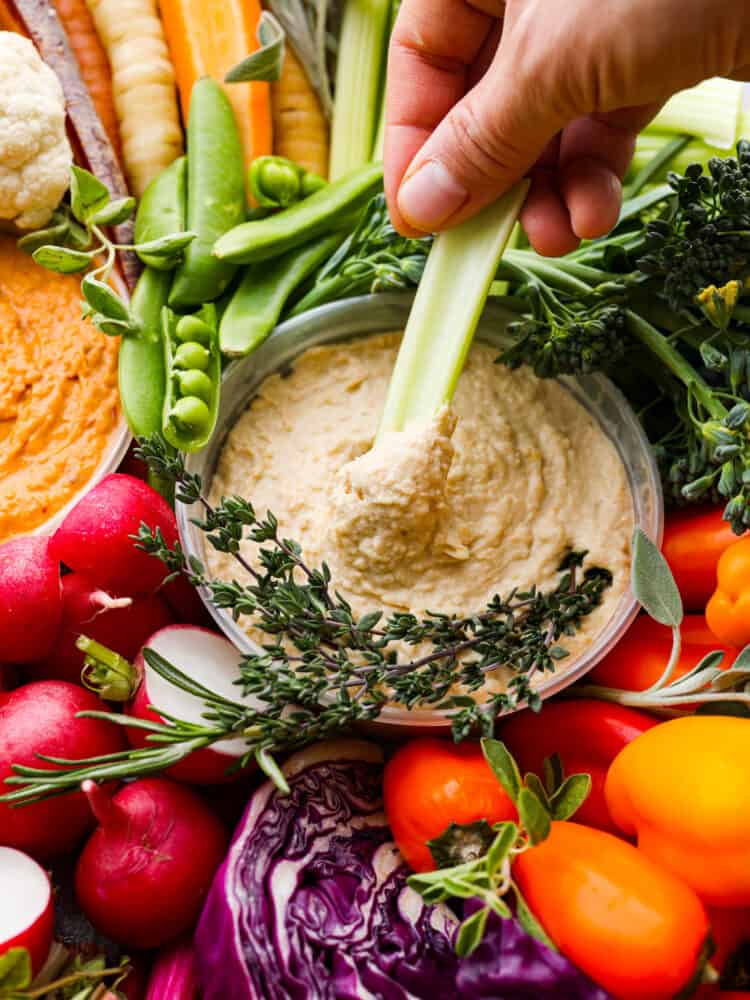 A hand dipping a celery stick into hummus from a crudités tray.