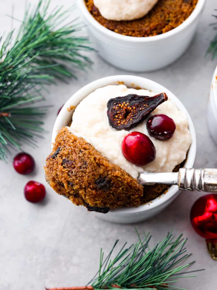 Top view of figgy pudding in a ramekin garnished with brandy butter and figs and cranberries on top. A spoon is lifting out a spoonful of pudding. Greenery and ornaments are scattered around the pudding.