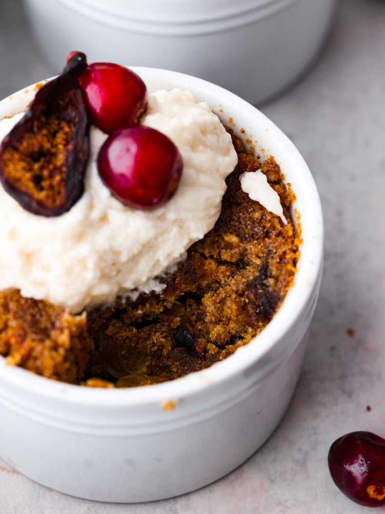 Close up view of figgy pudding in a ramekin garnished with brandy butter and figs and cranberries on top. A spoonful of pudding is missing from the ramekin.