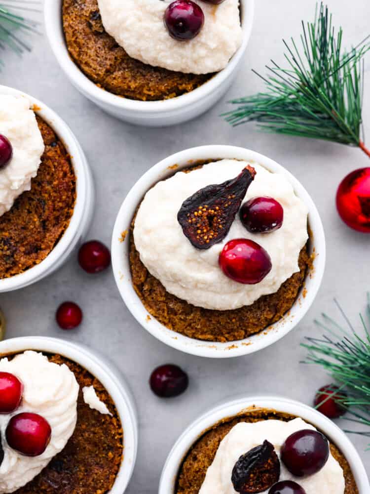 Top view of figgy pudding in ramekins garnished with brandy butter and figs and cranberries on top. Greenery and ornaments are scattered around the pudding.