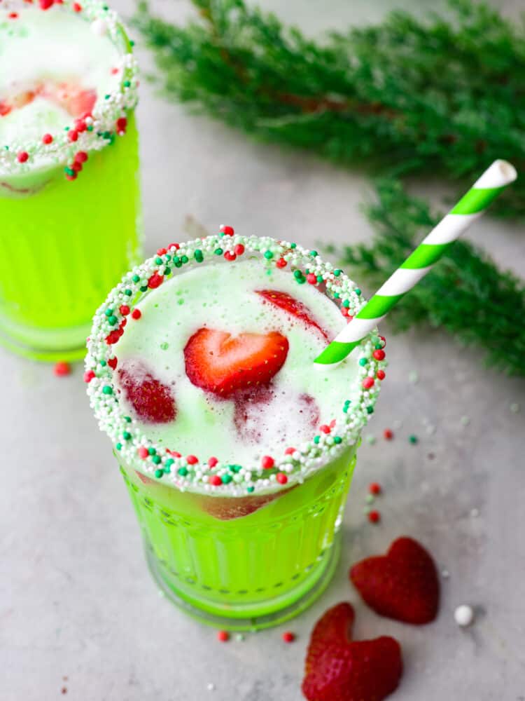 Top-down view of Grinch punch served in a glass, garnished with strawberries and sprinkles around the rim. There is a green and white swirled straw in the glass.