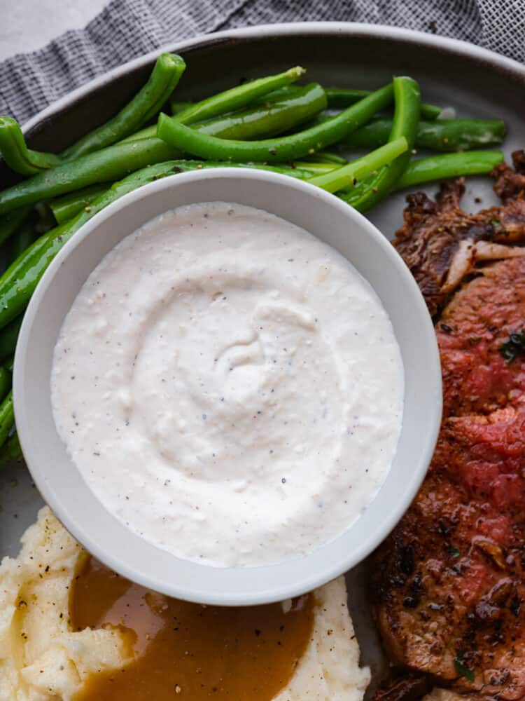 Horseradish sauce in a bowl with green beans, mashed potatoes, and a steak on a plate.