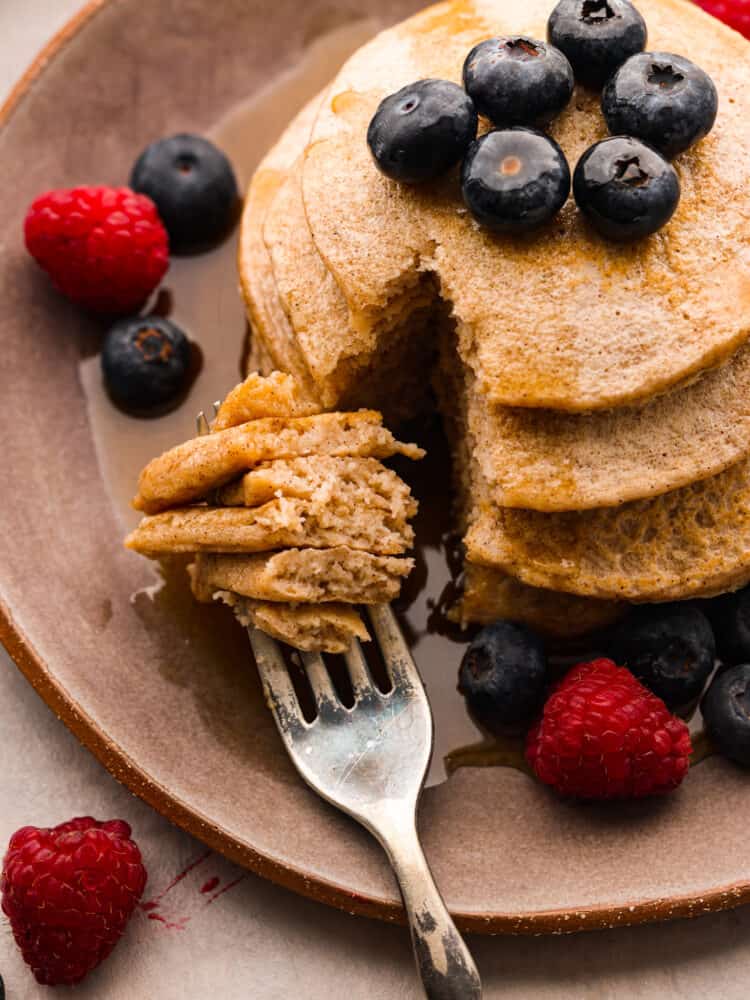 Close up overhead view of protein pancakes on a brown plate garnished with fresh blueberries, raspberries and maple syrup. A fork is in the plate, making cuts in the pancake.