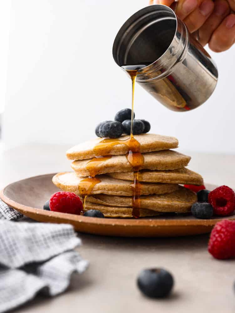 Close view of maple syrup being poured onto the protein pancakes.  The pancakes are on a brown plate garnished with fresh blueberries and raspberries.