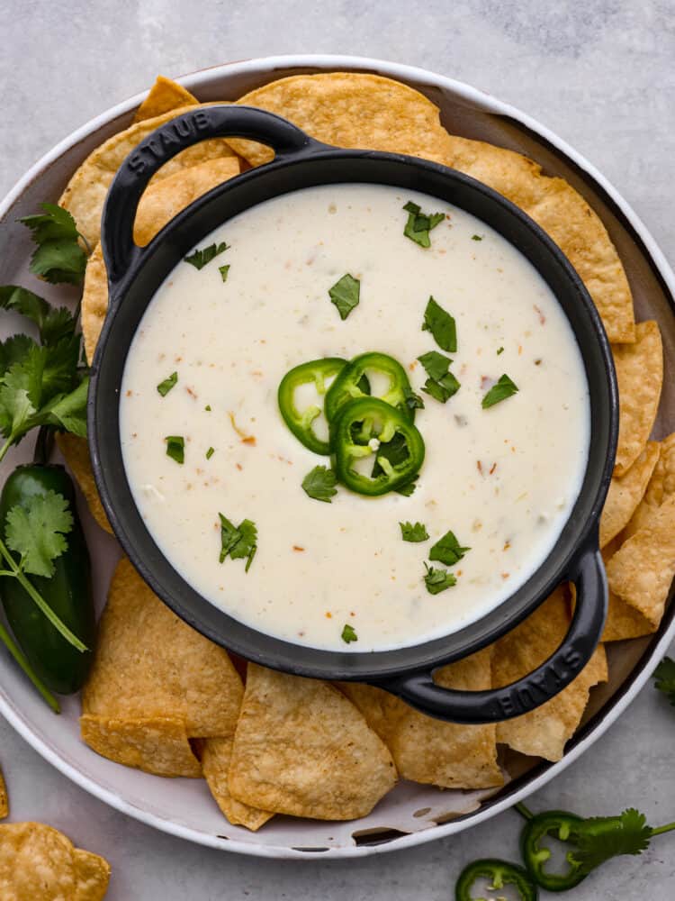 Queso blanco dip in a black bowl with fresh jalapenos and chips around it.
