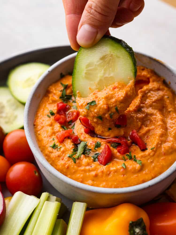 Red pepper hummus in a bowl with a cucumber being dipped in it.