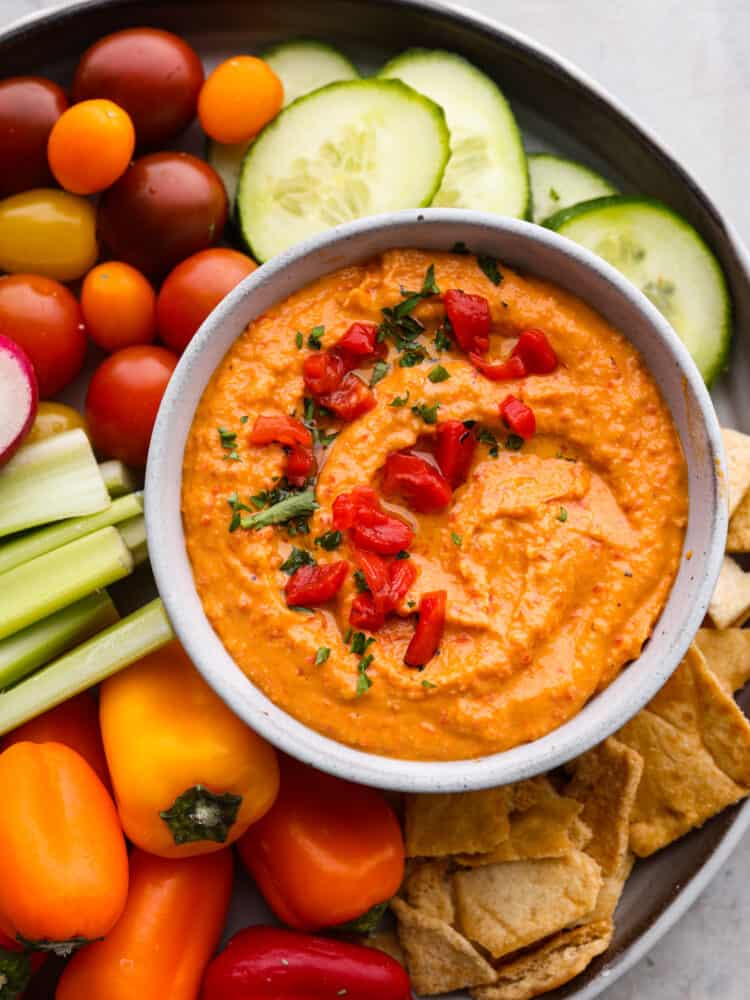 Roasted red pepper hummus in a bowl with fresh veggies around it.