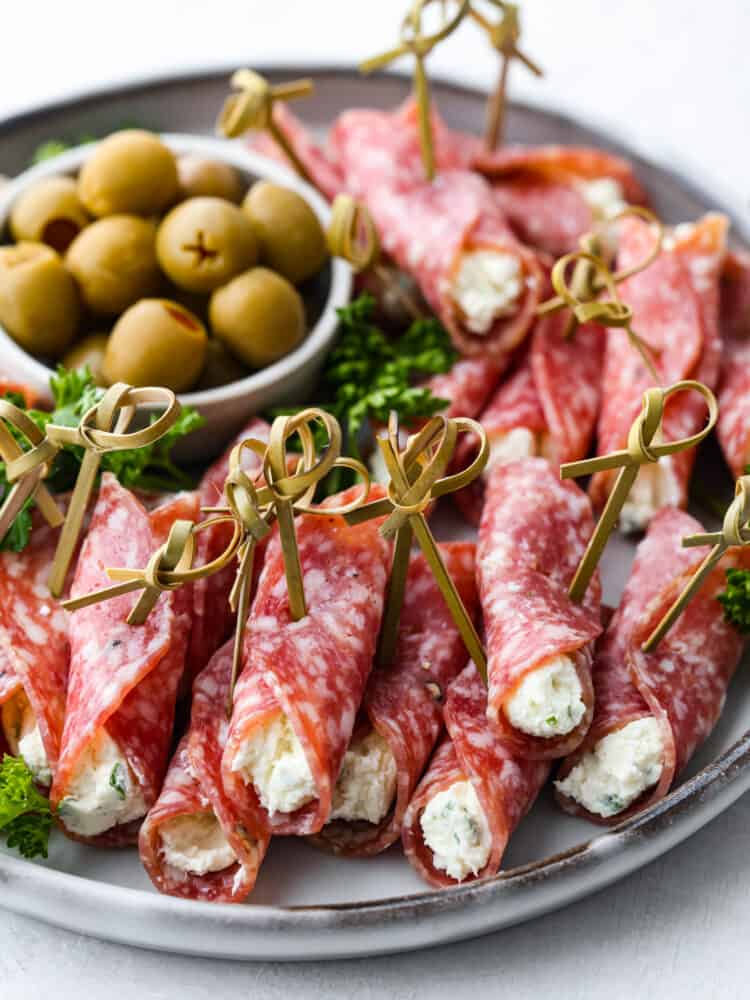 Salami roll ups on a plate with a bowl of olives in the background.