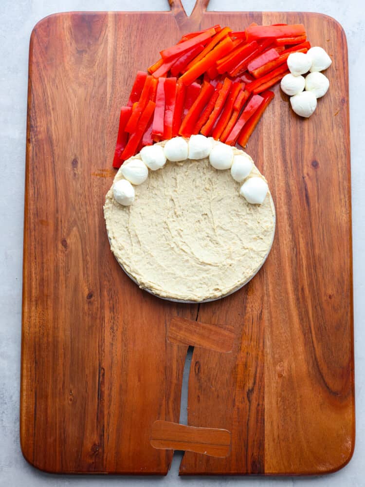 Hummus, mozzarella cheese balls, and sliced red peppers being shaped to look like a face and Santa hat.