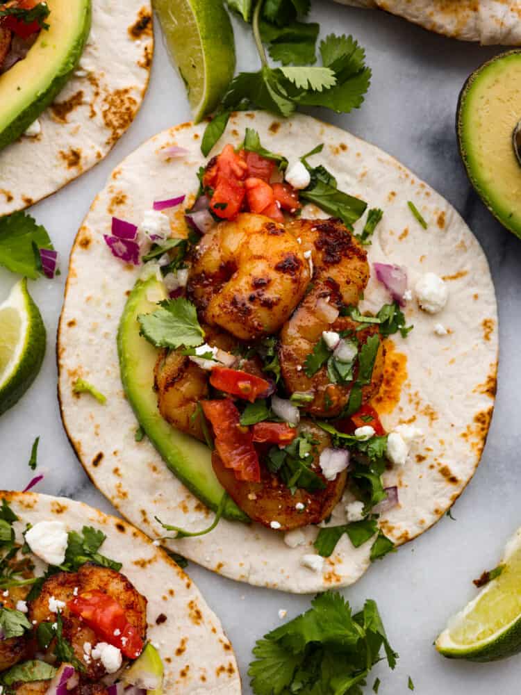 A flour tortilla topped with shrimp, sliced avocados, tomatoes, red onion, and cilantro.