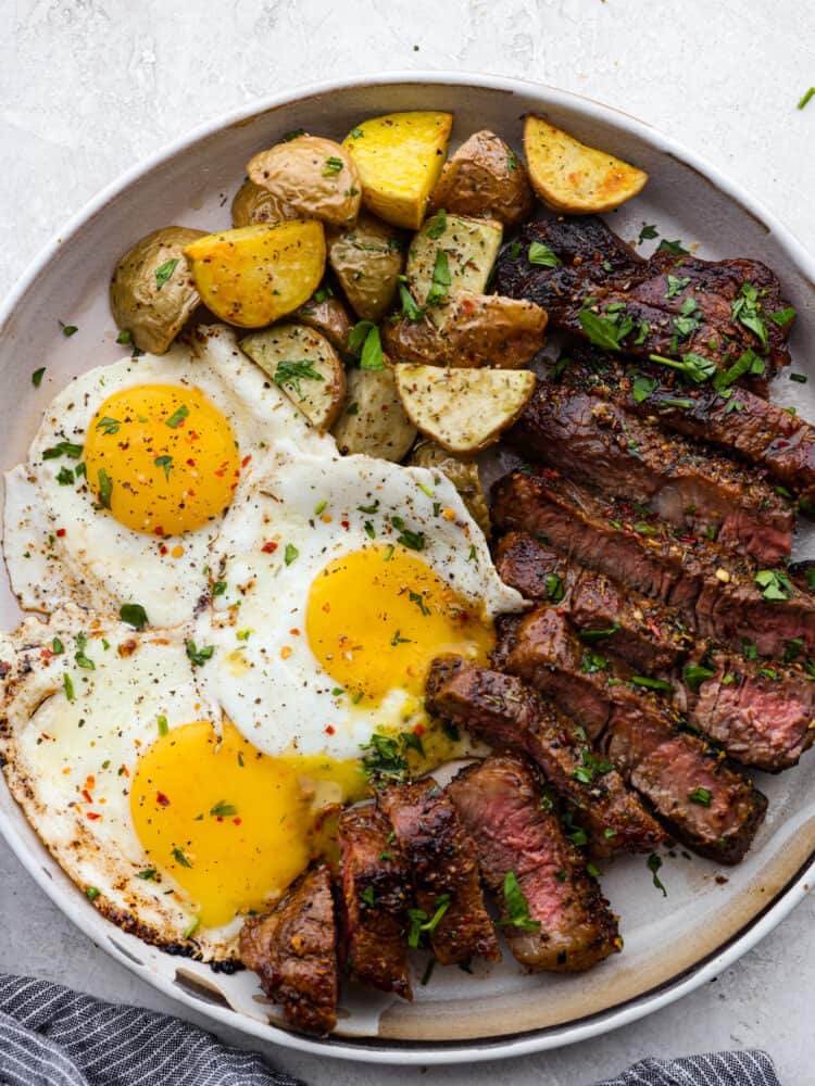 Top view of steak and eggs on a gray plate with a side of roasted potatoes. Garnished with chopped parsley.