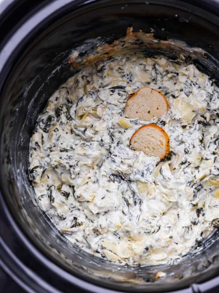 Spinach artichoke dip in a slow cooker with two pieces of crostini in it.