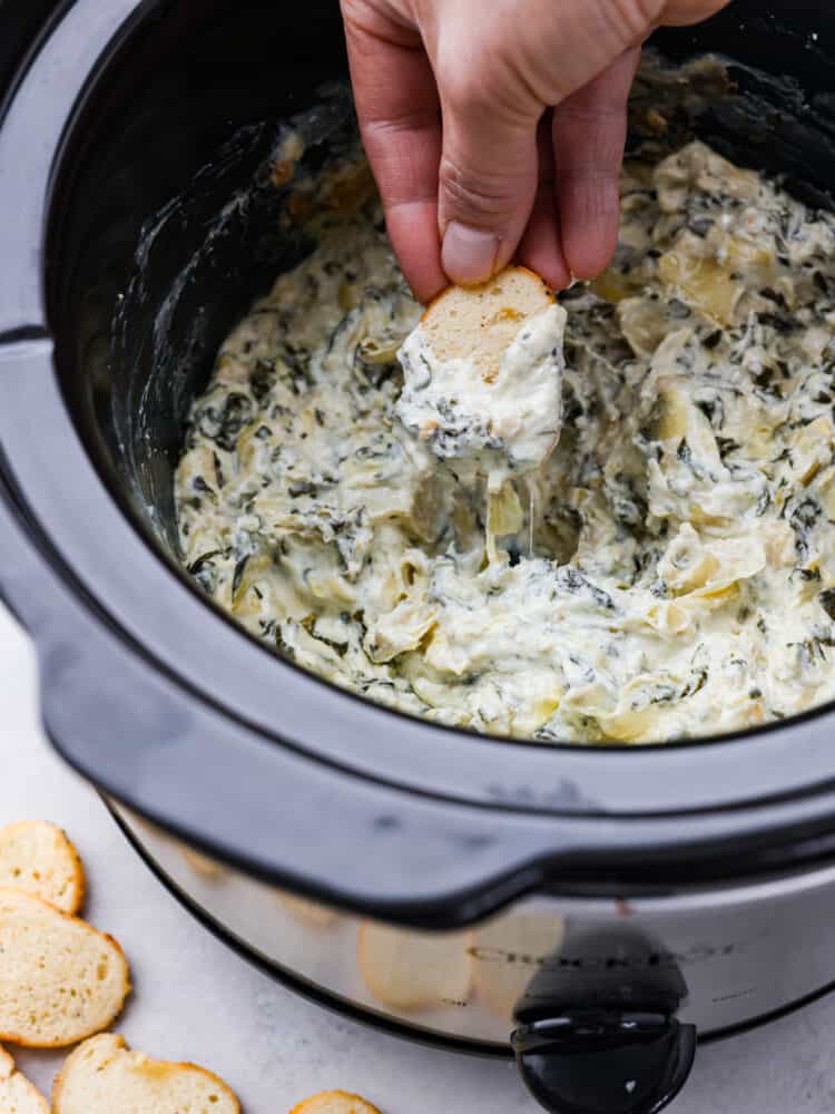 Crostini being dipped into a a slow cooker filled with spinach artichoke dip.