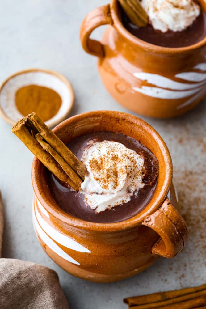 Mexican hot chocolate served in a tan colored mug, topped with a cinnamon stick.