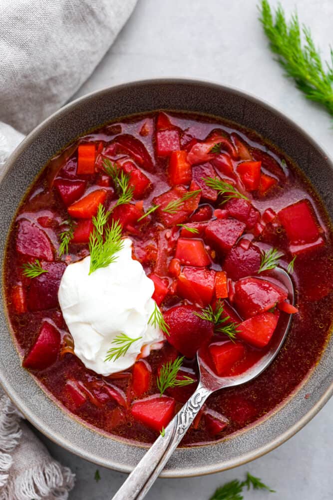 Beet soup served in a gray bowl with a dollop of sour cream and some fresh dill on top.