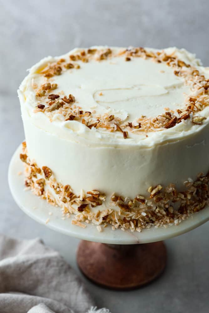 A whole Italian cream cake on a stand topped with chopped pecans and coconut.