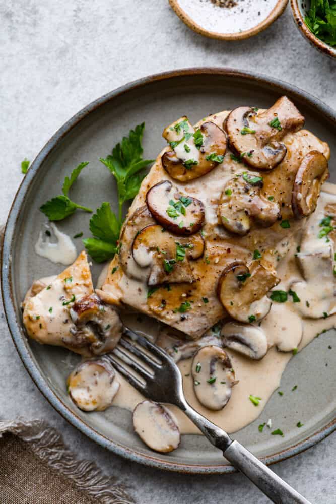 Creamy Chicken Marsala is a classic Italian dish that is ready in under 30 minutes! Marsala wine is made into a creamy sauce paired with mushrooms for the ultimate savory bite. It's simple and easy to whip up and sure to be a crowd-pleaser! A piece of chicken on a plate with a fork taking a bite out of it.