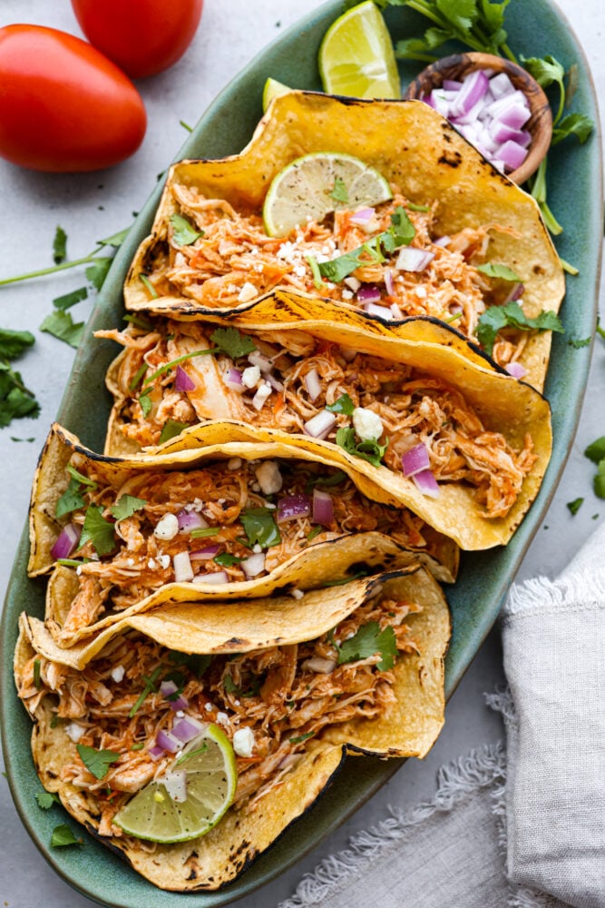 Top-down view of 4 chicken tacos on a green plate. They are topped with chopped onions, cheese crumbles, and cilantro.