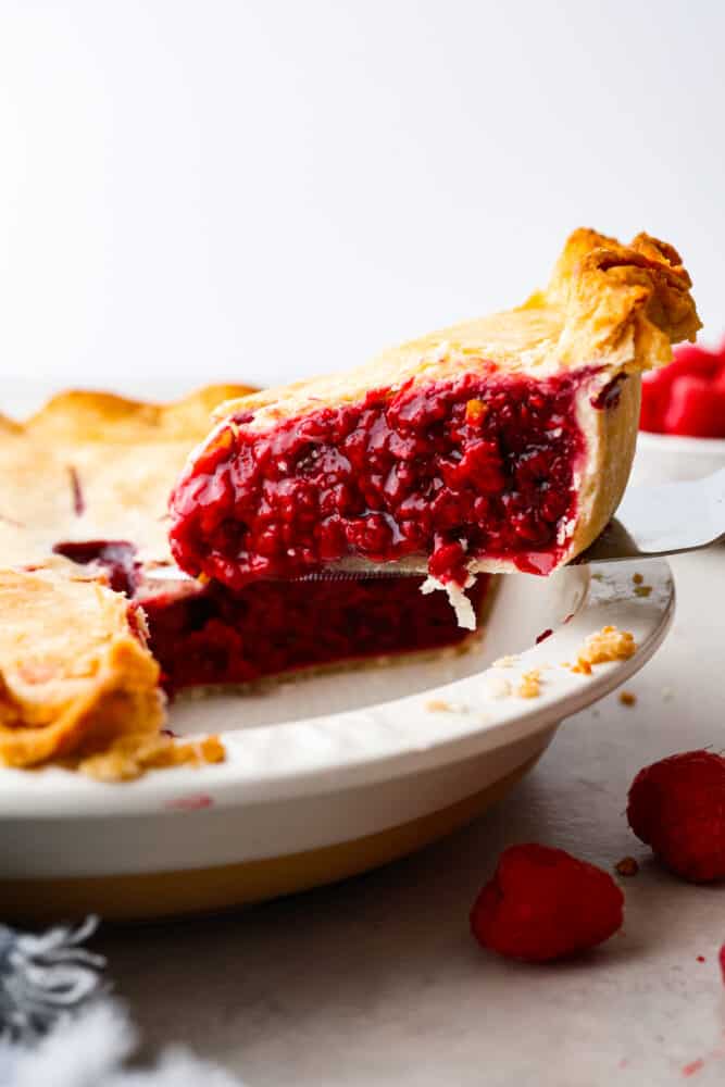 Raspberry pie is a timeless dessert that has been enjoyed for generations, and for good reason. With its flaky, buttery crust and tangy-sweet filling bursting with fresh, juicy raspberries, it’s the perfect balance of flavors and textures. And while store-bought options are convenient, nothing beats making it from scratch! A slice of pie being served with a cake spatula.