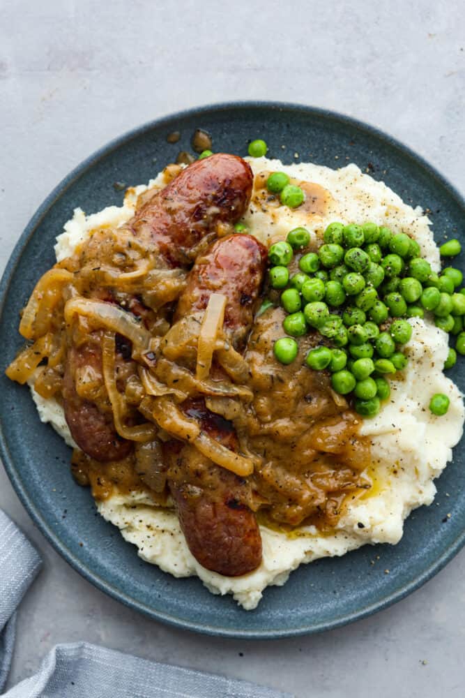A gray plate topped with mashed potatoes, 2 sausages, onion gravy, and peas.