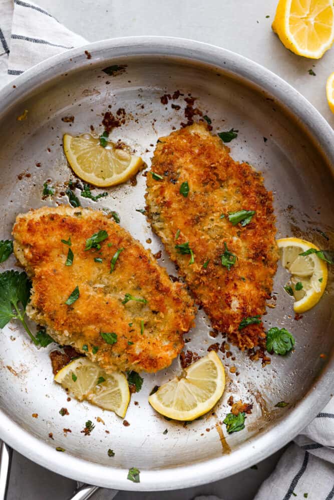 Top view of chicken Milanese in a silver skillet. Chopped parsley and lemon slices are garnished on top of the chicken.