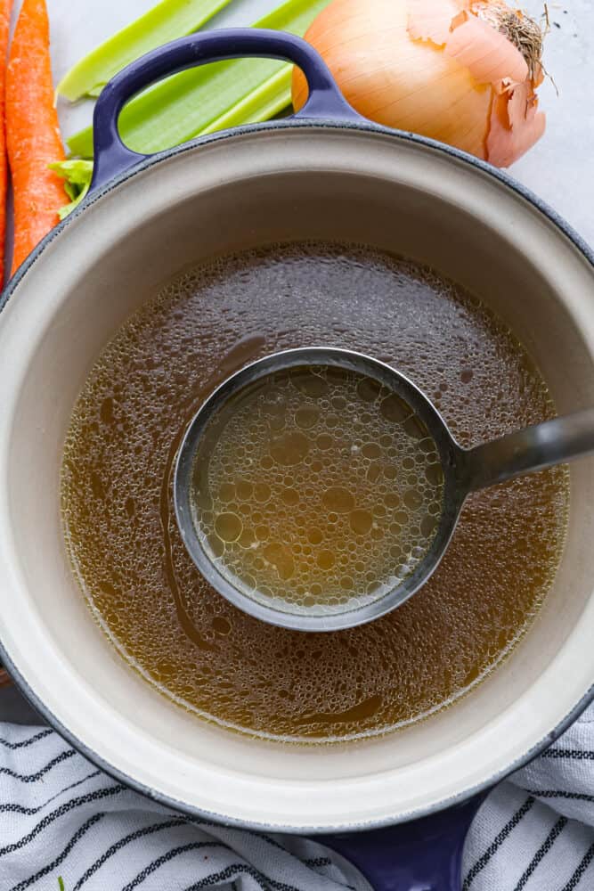 Top view of a ladle lifting up chicken broth from a blue pot. Vegetables and a towel are scattered on the counter around the pot.