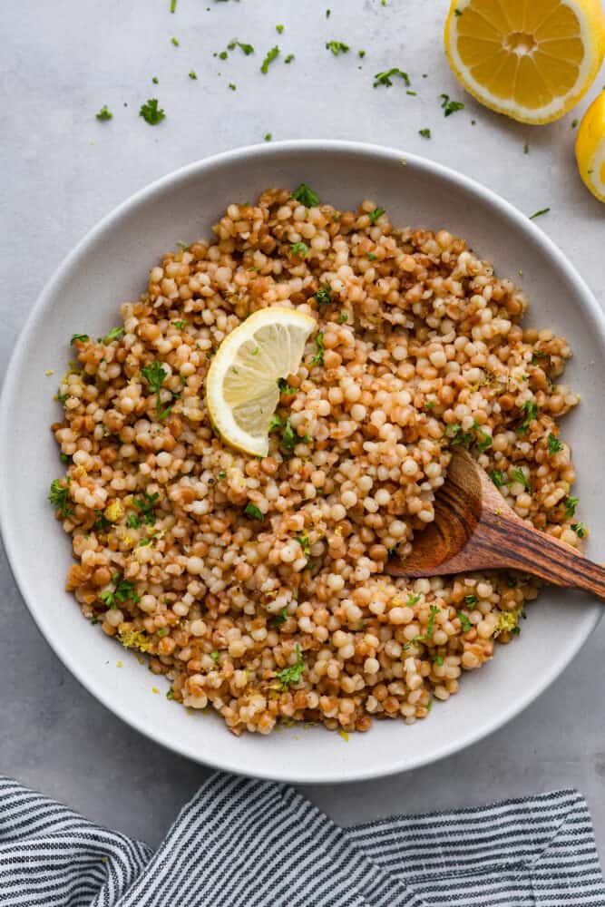 Looking to add a burst of Mediterranean flavor to your dining table? This delicious Israeli couscous is the answer! With its aromatic blend of olive oil, lemon zest, garlic, and fresh parsley, you won't be able to get enough. It's the perfect side dish for summer! Top-down view of couscous in a white bowl, garnished with parsley and a lemon wedge.