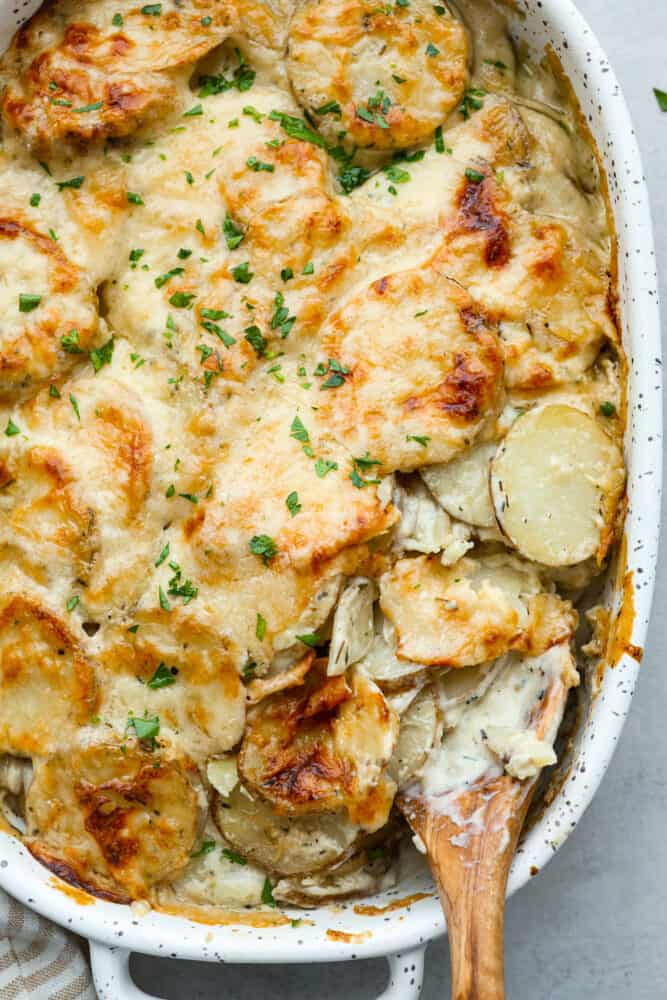 Hero image of a scoop of scalloped potatoes being taken out of a white baking dish.