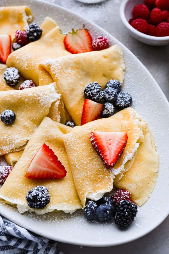 Close up of folding crepes on a gray platter. The crepes are dusted with powdered sugar and topped with berries.