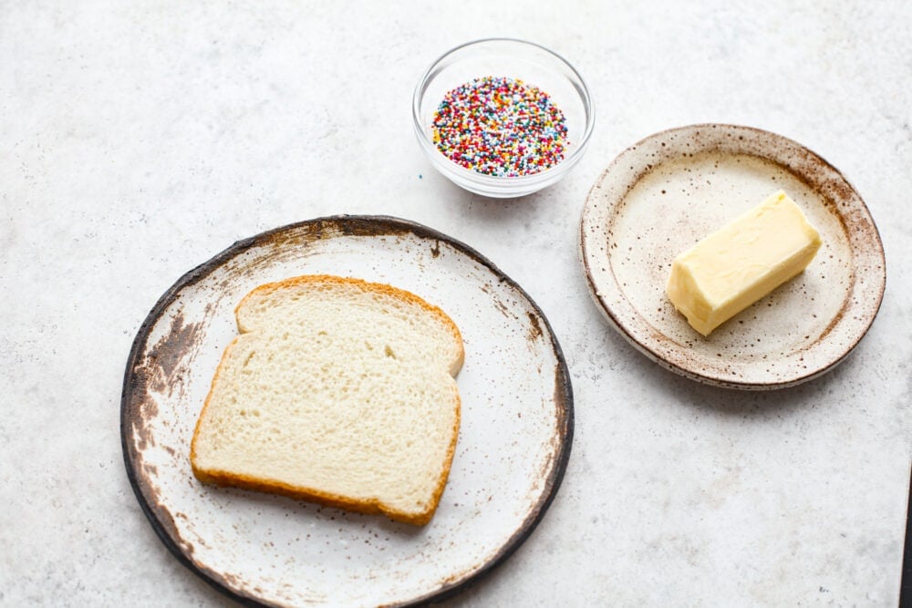 A plate of bread, a bowl of sprinkles, and a stick of butter.