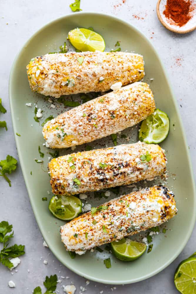 Grilled Mexican Street Corn is the perfect grilled corn slathered with an amazing mayonnaise blend and topped with parmesan cheese and chili powder. This is one of the best corn recipes you will eat! A sage green platter with 4 ears of corn slathered with the mayonnaise mixture and sprinkles with spices and cheese. Lemon wedges garnish the plate.