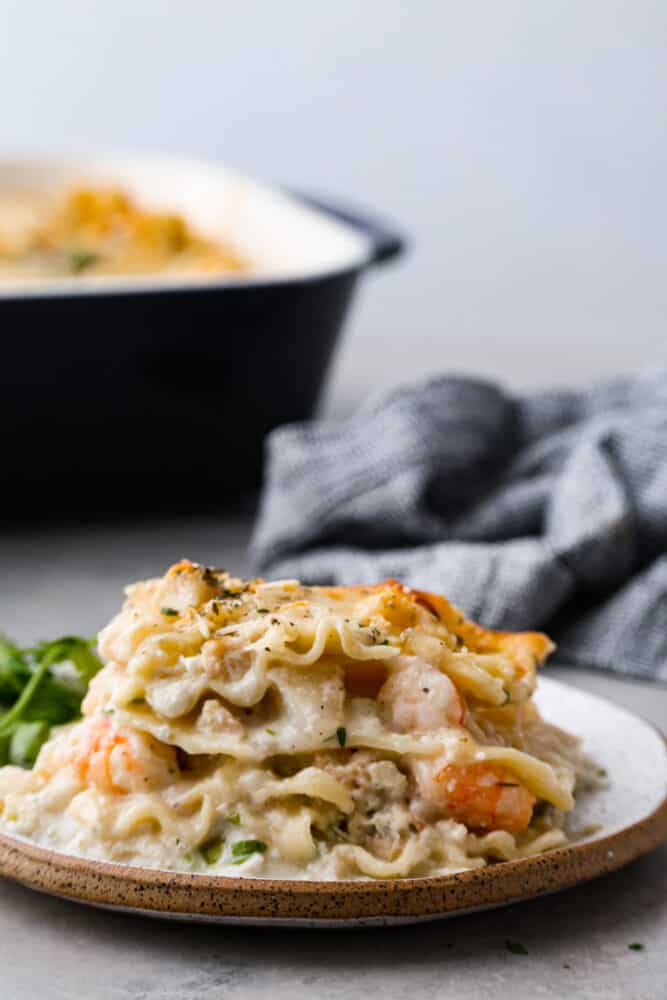 Close up view of seafood lasagna on a white plate with the pan of lasagna is in the background.