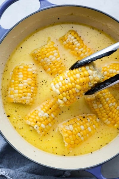 This is the BEST recipe for how to boil corn on the cob! Fresh summer corn gets boiled in a sweet creamy mixture of milk, butter, sugar, and salt. Cook and flavor your corn all in one pot! Close top view of tongs lifting up corn from a pot.