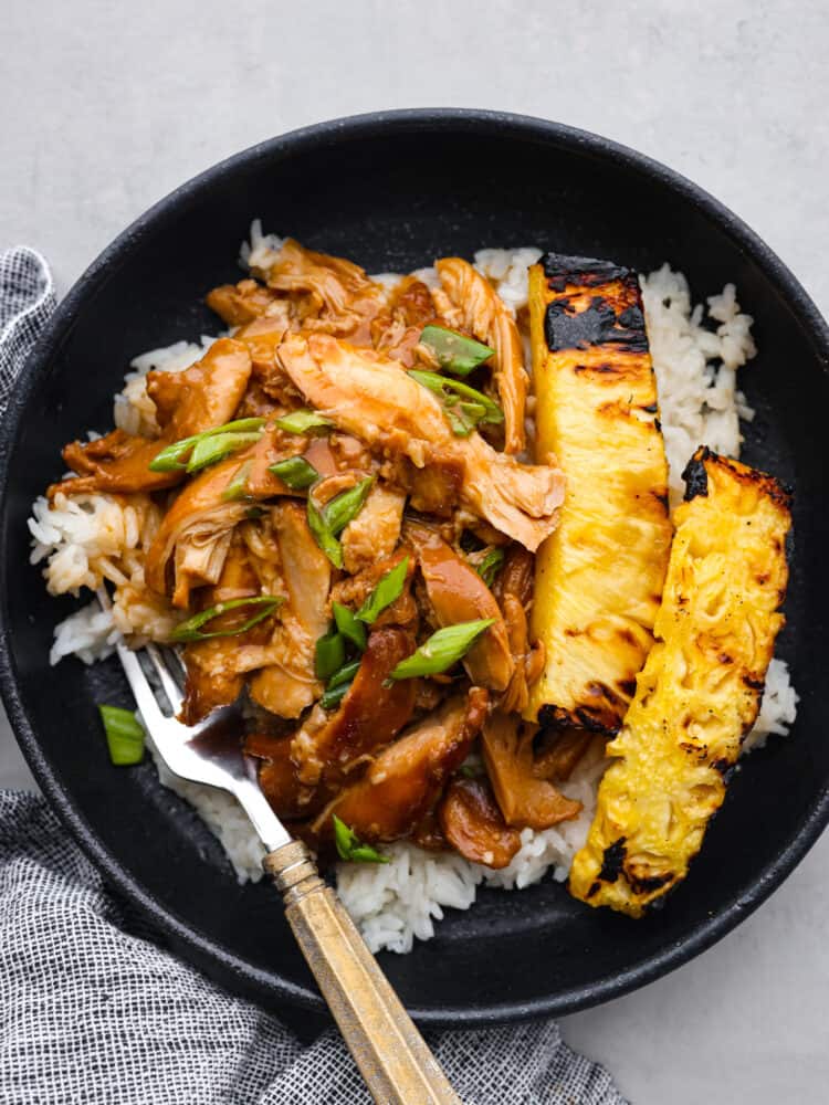 Huli huli chicken served in a black bowl along with some white rice and grilled pineapple.