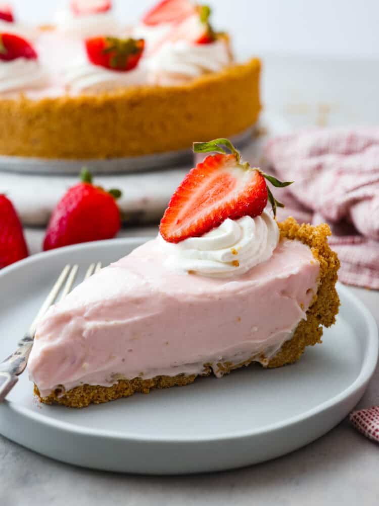 A close up view of a slice of no-bake strawberry cheesecake on a light blue colored plate.The pie is garnished with whipped cream and a strawberry slice. A fork is on the plate next to the slice.