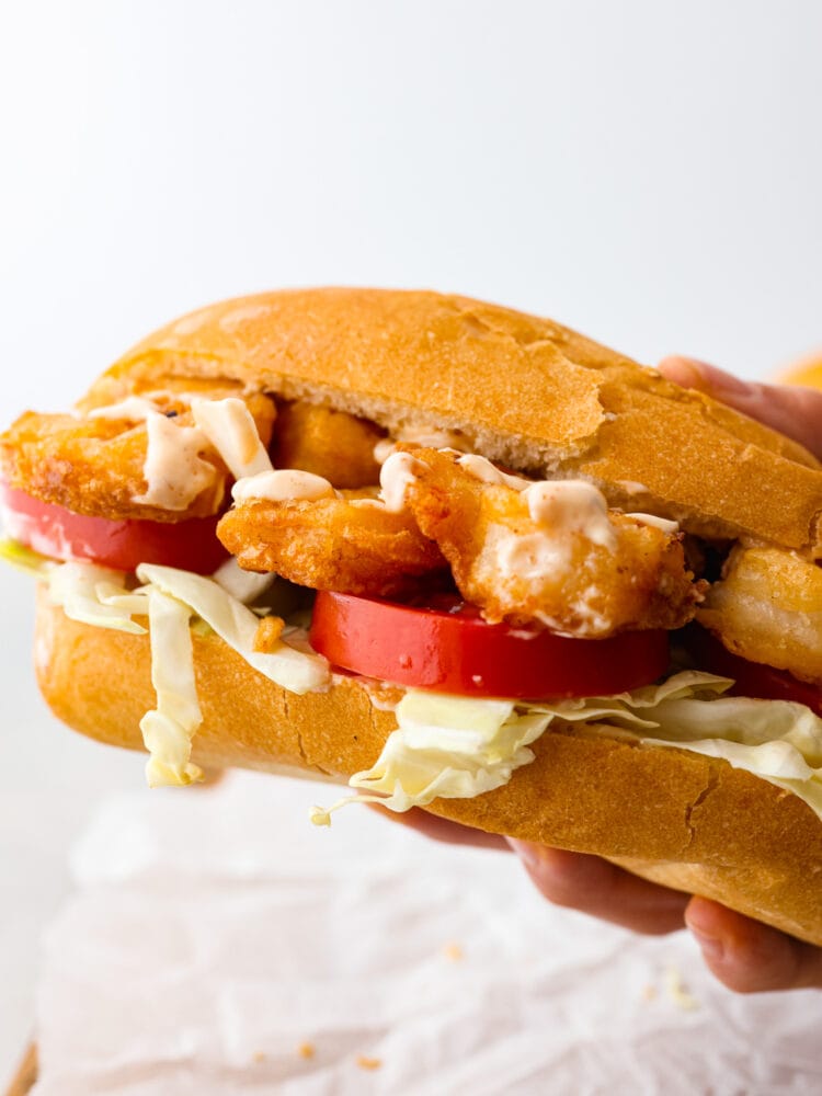 Close up view of a shrimp po boy sandwich being held up. Crispy fried shrimp, tomatoes, and cabbage are nestled in French bread.