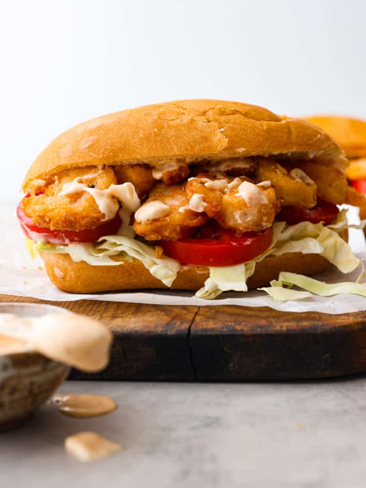 Close up view of shrimp po boy on a wood board. Cajun aioli is in a bowl next to the sandwich with the sauce dripping off the knife.