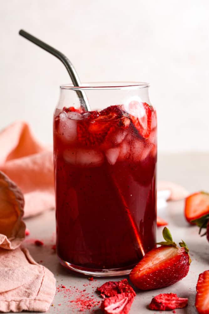 The side view of a strawberry acai refresher in a clear cup with a metal straw and strawberries scattered around the cup. 