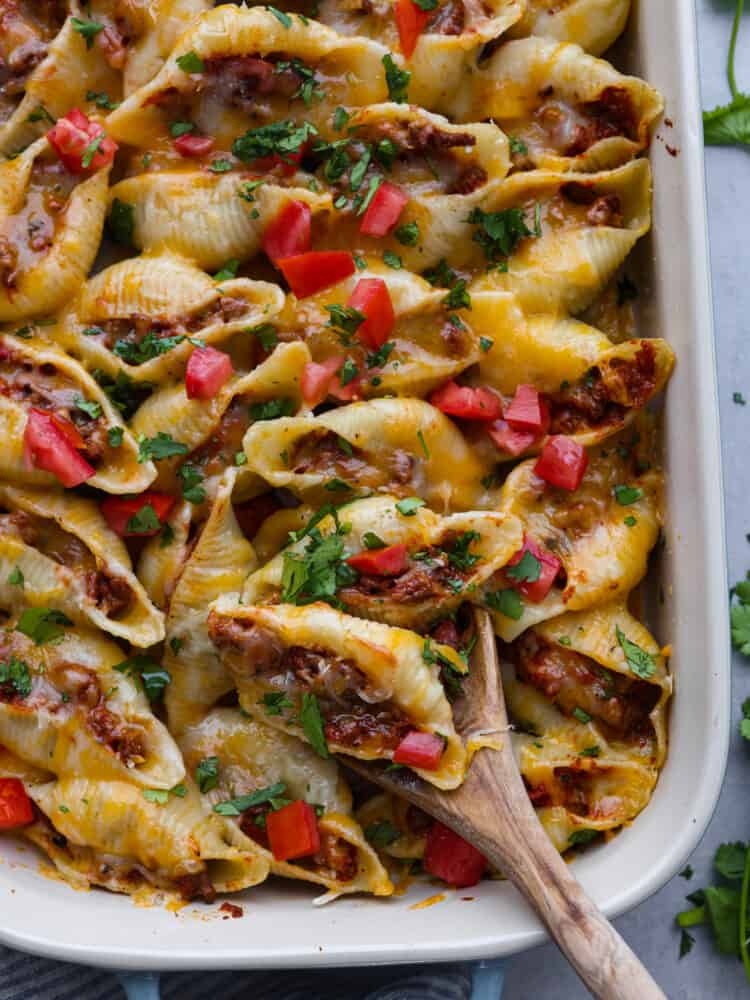 Top view of taco stuffed shells in a baking dish and garnished with diced cilantro and tomatoes. A wooden spoon is lifting up some cooked shells. 