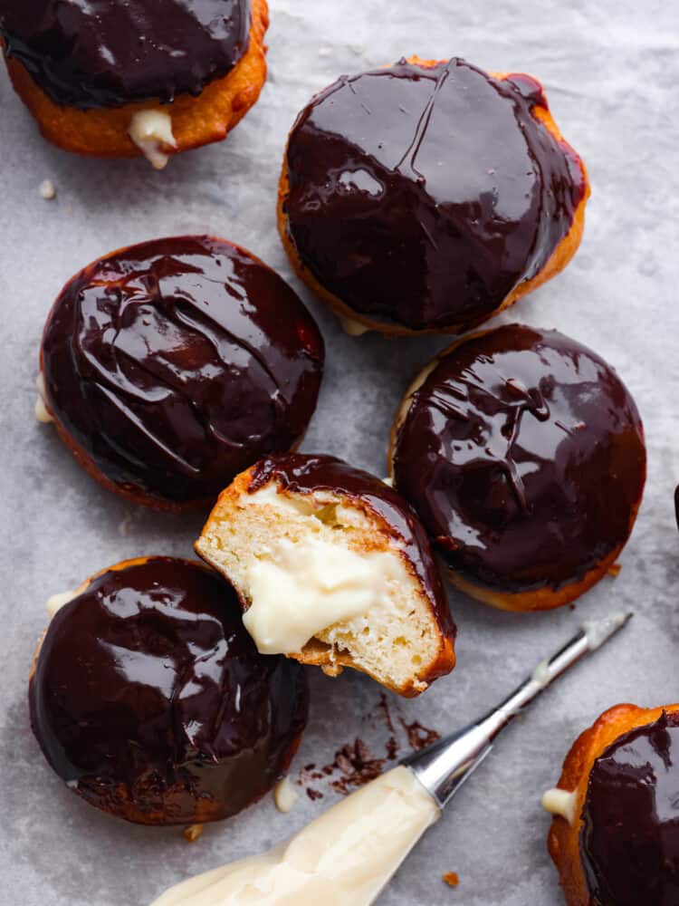 Treat yourself to a batch of homemade Boston Cream Donuts covered in chocolate ganache. These delightful treats are made from scratch and create an irresistible combination of fluffy donuts, creamy filling, and delicious chocolate topping. The top view of Boston cream donuts on parchment paper. One is cut open so you can see the cream inside.