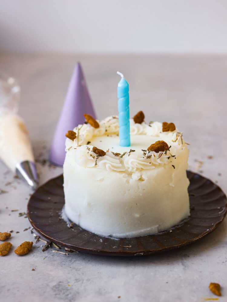 A cat birthday cake with a blue candle on it. There is a purple party hat in the background.