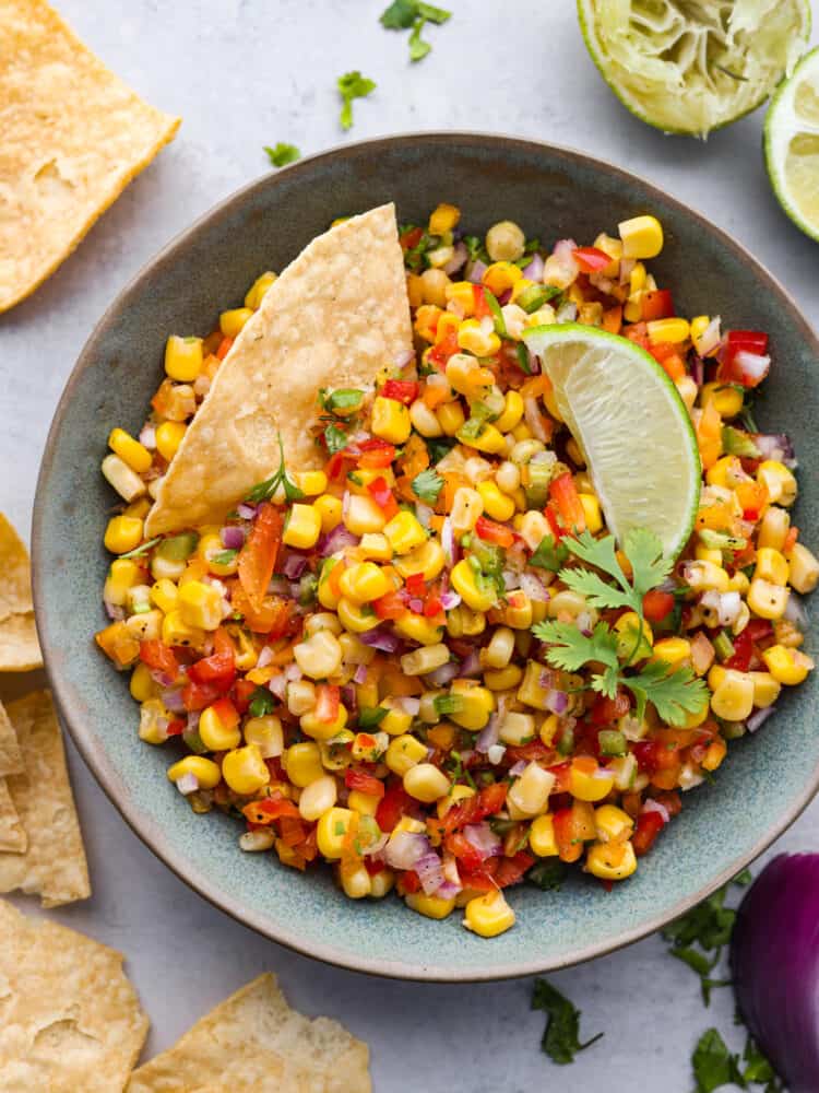 The top view of a bowl of corn salsa garnished with cilantro and topped with a lime and a tortilla chip. 